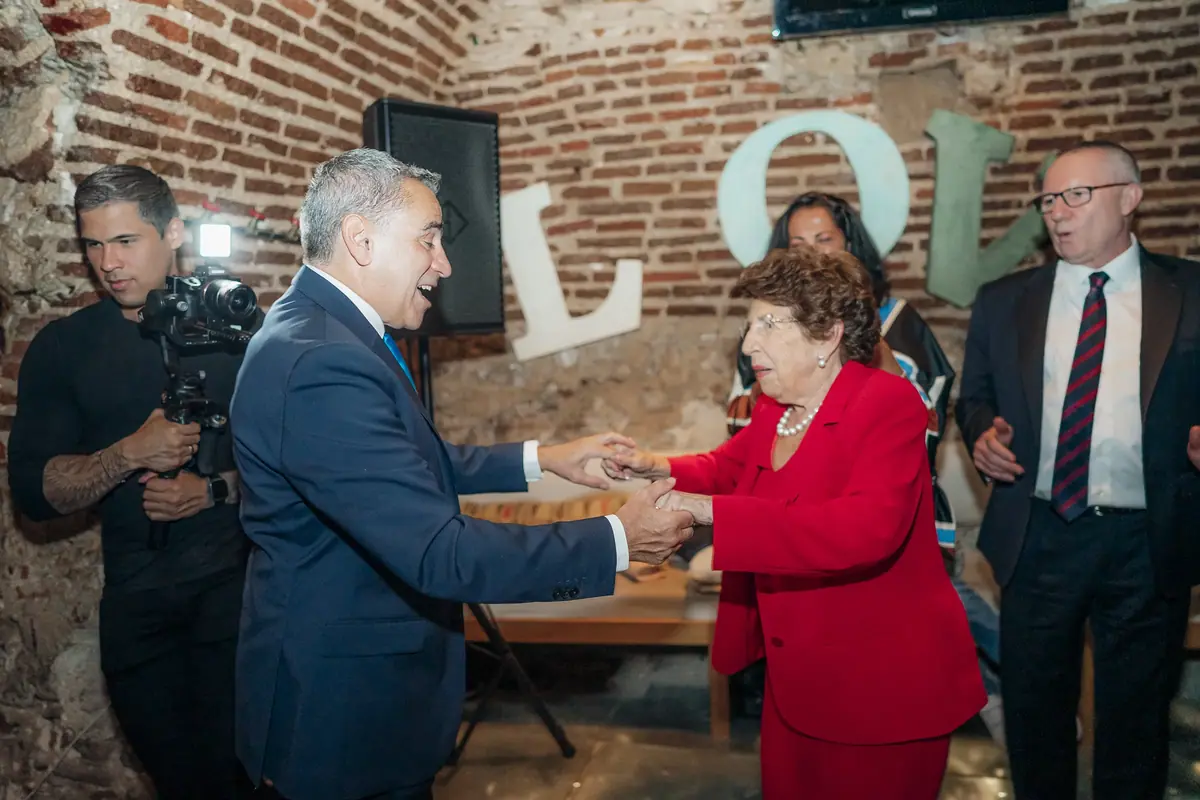 A man in a navy suit and an older woman in red, wearing a pearl necklace, clasp hands in greeting against exposed brick, with guests around them and a camera operator on the left
