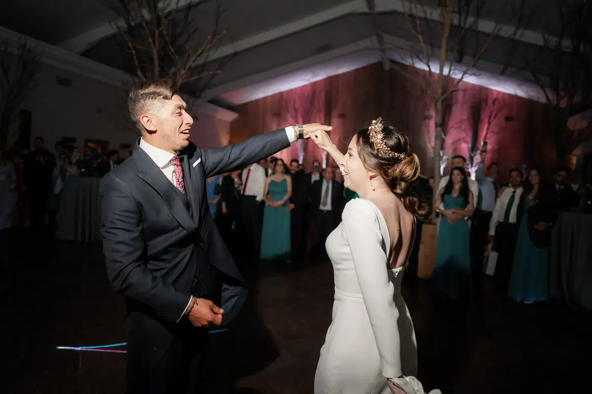 Los novios bailan en una sala en penumbra, con las paredes iluminadas en tonos rosados y los invitados mirando al fondo