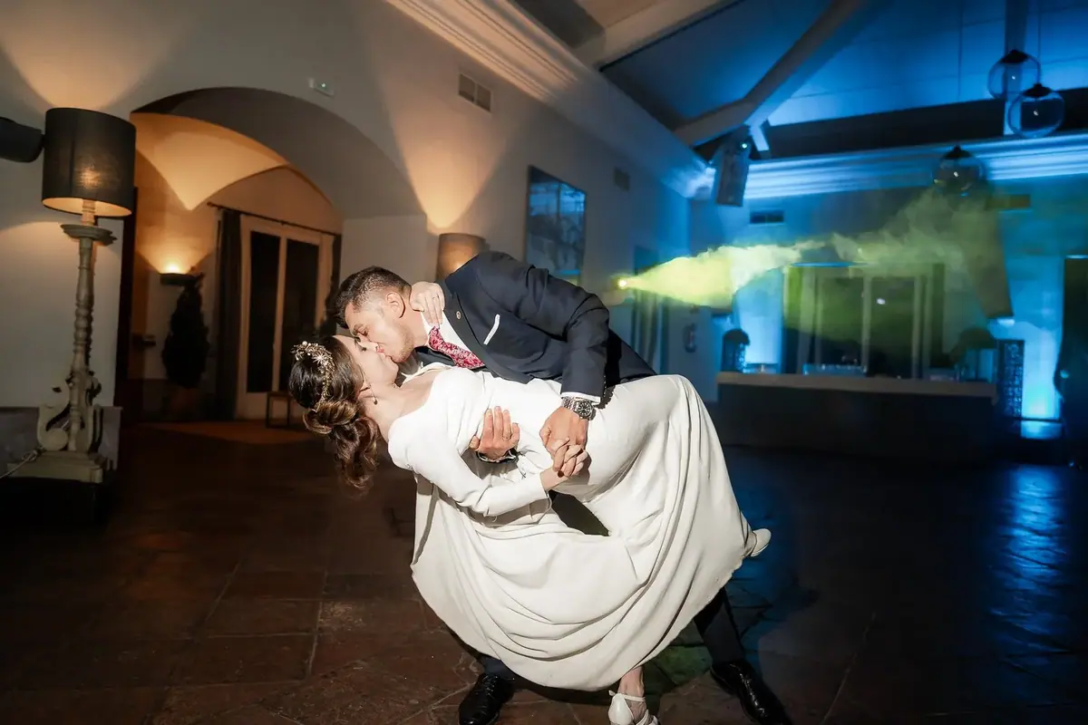 El novio inclina a la novia para besarla en el baile, con luces azules y algo de humo al fondo de la sala