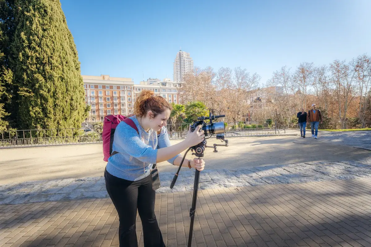 A videographer films with a camera on a tripod on a tree-lined promenade, under a blue sky