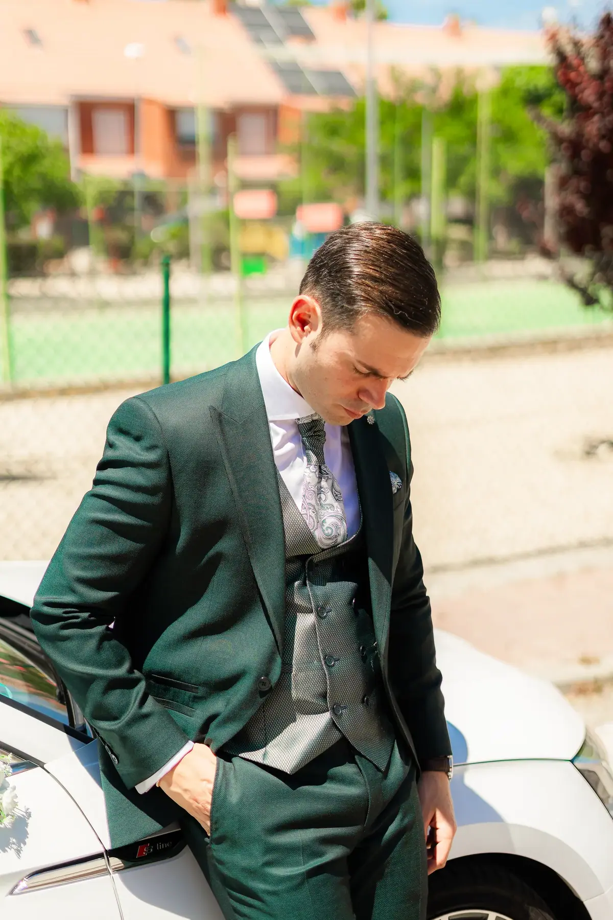 El novio, con traje verde, de pie junto a un coche y con la mirada baja, a solas antes de la ceremonia