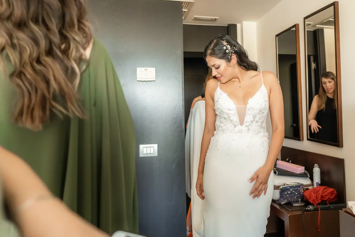 The bride, in a strappy dress with her hair up, looks down as she stands beside some mirrors, while a person in the foreground, seen from behind, fills half the frame