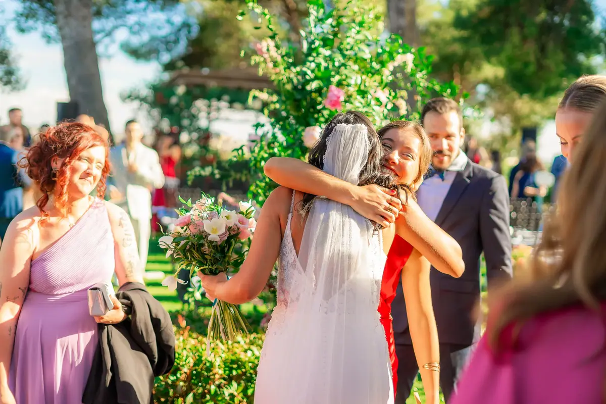 The bride, in a veil and holding her bouquet, hugs a guest in red in a garden, among other smiling guests