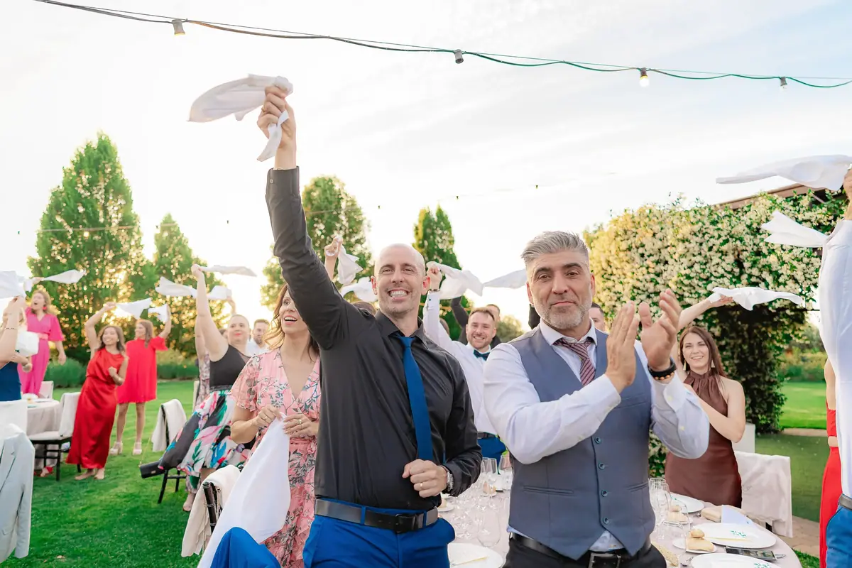 Invitados en un jardín, bajo una guirnalda de luces, agitan servilletas blancas y aplauden con la hierba y los árboles al fondo