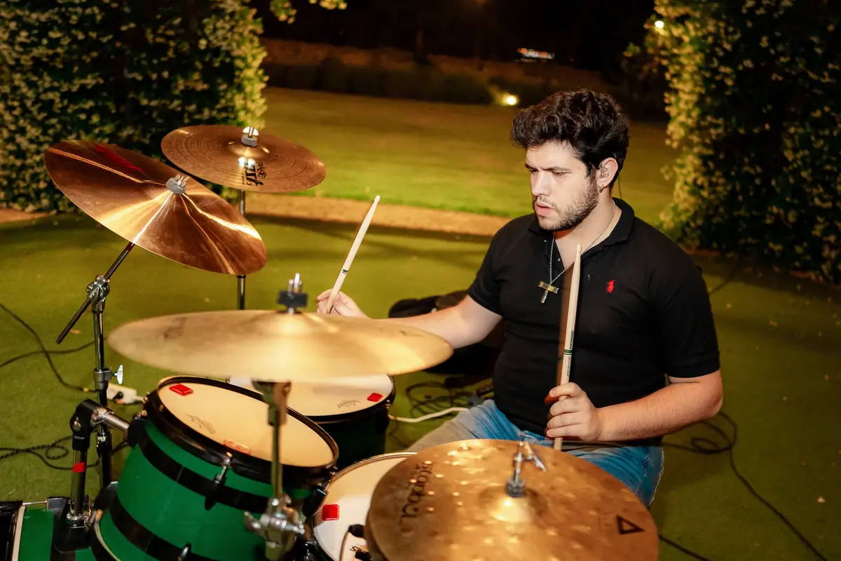 A drummer plays at night in a garden, with the lawn lit and hedges behind