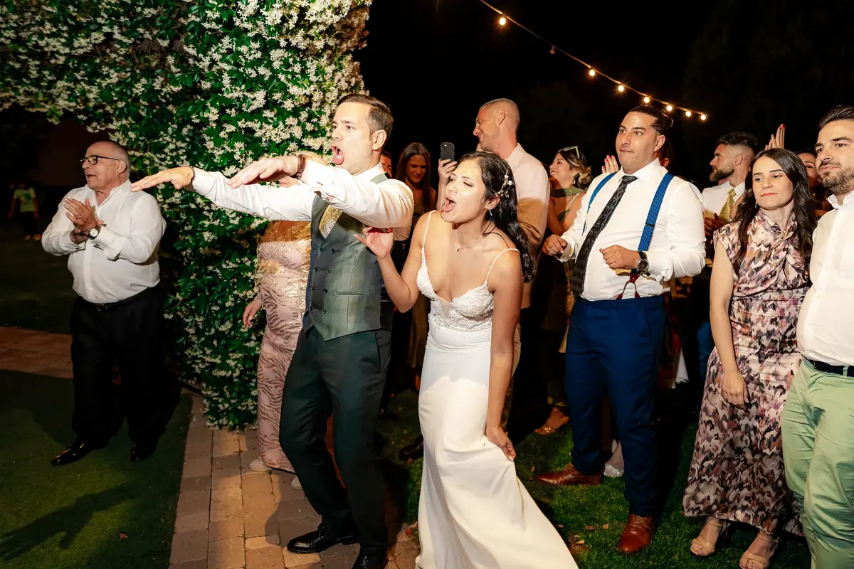 La novia, de vestido blanco, baila de noche en un jardín junto al novio, con los invitados alrededor bajo una guirnalda de luces