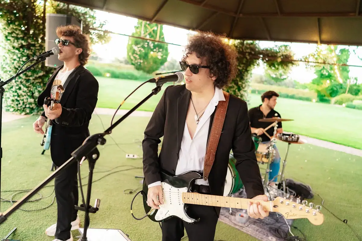 Two musicians with guitars and sunglasses play at their microphones on a lawn in a garden, with a drummer behind them