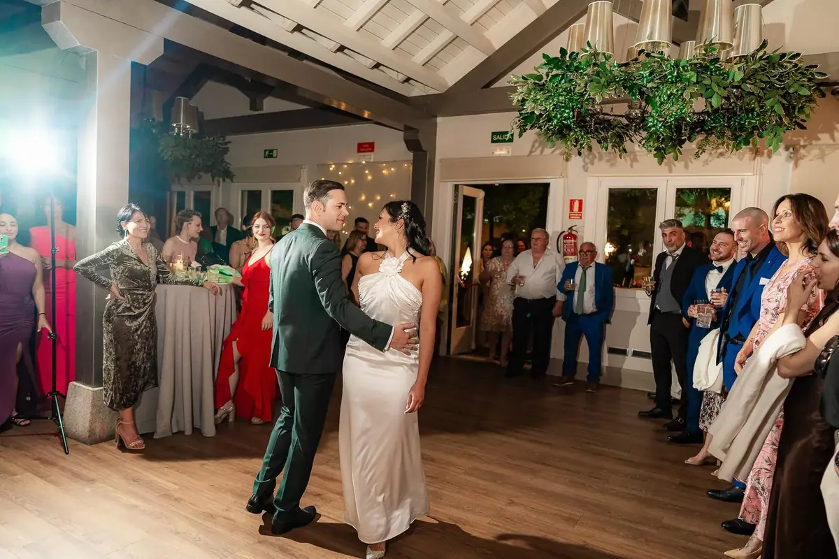 Los novios bailan en un salón con vigas de madera y una lámpara de ramas verdes, rodeados de invitados que los miran en círculo