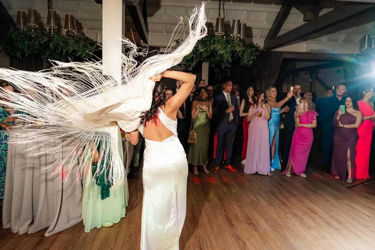 A woman in a white dress dances with her back to us, a white fringed shawl flying in the air, in front of guests who watch and clap in a room with dark beams